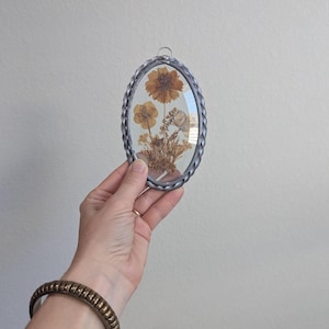May include: Oval-shaped pressed flower art piece with a silver-colored braided metal frame. The artwork features dried flowers in shades of brown and yellow, encased in glass. A small metal loop is at the top for hanging.