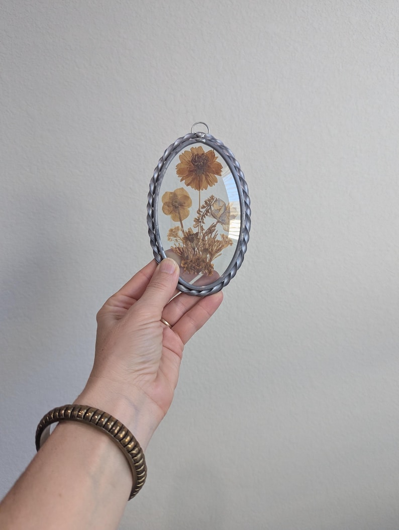 May include: Oval-shaped pressed flower art piece with a silver-colored braided metal frame. The artwork features dried flowers in shades of brown and yellow, encased in glass. A small metal loop is at the top for hanging.