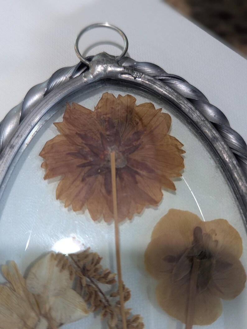 May include: Oval-shaped, silver-toned framed pressed flower art. The frame has a twisted rope design. Inside, dried flowers in shades of brown and beige are visible, with a clear glass cover and a metal loop for hanging.