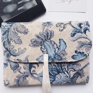 Puede incluir: Un bolso de mano con estampado floral en tonos crema y azul. El bolso tiene un cierre de solapa y una cinta color crema. El diseño incluye un patrón floral con hojas y flores azules. También se ve una tarjeta de visita con el texto "ATELIER DAILY ELEGANCE".