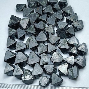 Pode incluir: Uma coleção de cristais escuros e geométricos com um brilho metálico. Os cristais são de uma cor cinza-esverdeada escura e têm uma superfície áspera e texturizada. Estão dispostos sobre uma superfície branca, com uma régua em primeiro plano.