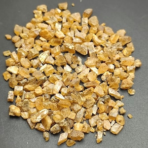 May include: A pile of small, rough-cut, amber-colored gemstones. The crystals vary in size and shape, with a translucent quality. The stones are scattered on a dark surface, creating a contrast that highlights their color and texture.