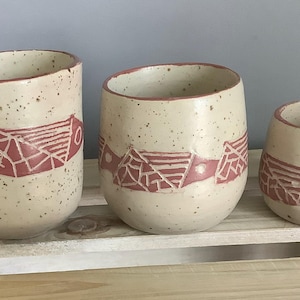 Op de afbeelding: Drie gespikkelde keramische bekers van verschillende formaten. Elke beker heeft een crèmekleurige basis met een rood geometrisch ontwerp in het midden. De bekers worden op een houten oppervlak weergegeven.
