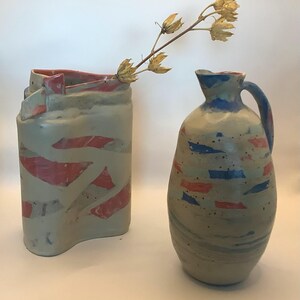 Peut inclure: Deux vases en céramique. L'un est un vase rectangulaire et haut avec des accents rouges et gris. L'autre est une cruche arrondie avec une anse, avec des détails bleus et rouges. Une tige de fleur séchée est placée dans le grand vase.