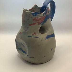Peut inclure: Pichet en céramique fait à la main avec une anse bleue et une forme irrégulière unique. Le pichet est principalement gris avec des éclaboussures de peinture bleue, rouge et verte. Il présente un trou sur le côté et une surface texturée.