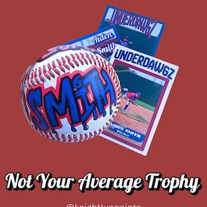 May include: A baseball with the name "SMITH" in blue graffiti-style lettering. The baseball is white with red stitching. Two baseball cards with the text "UNDERDAWGZ" and a player image are behind the ball. The text "Not Your Average Trophy" is below.