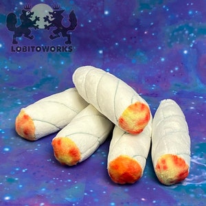 Può includere: Una collezione di oggetti cilindrici in peluche bianco con punte arancioni e gialle, simili a sigarette arrotolate. Ognuno ha un motivo a spirale cucito in azzurro. Lo sfondo è un motivo a tema galassia con il testo "LOBITOWORKS" e un logo.