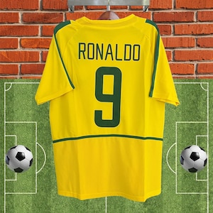 May include: A yellow football shirt with the name "RONALDO" and the number "9" printed in green. The shirt has green detailing on the sleeves and hem. The background shows a brick wall and a football pitch.
