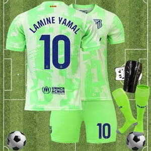 May include: A full football kit in green and white, with a jersey displaying "LAMINE YAMAL" and the number "10" in navy. The set includes shorts, socks, shin guards, and a football. The UNHCR ACNUR logo is also present on the jersey.