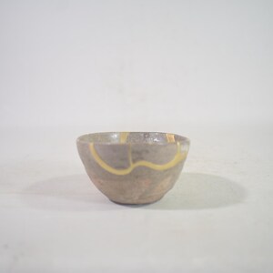 May include: A small, handcrafted ceramic bowl with a grey-brown exterior. The bowl features a unique design with gold-coloured accents that highlight the cracks and imperfections. The bowl is set against a plain white background.