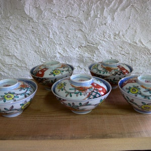 May include: Five ceramic bowls with lids, featuring a traditional Japanese design. The bowls are white with colorful floral and bird motifs, accented with red, green, and yellow. The lids have a small handle on top.