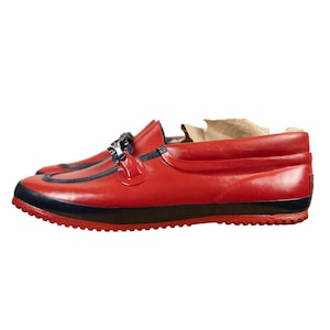 May include: A red slip-on loafer with a dark blue trim along the sole and vamp. The shoe features a decorative dark blue metal bit detail across the top and a textured rubber outsole. The interior contains a light brown paper insert.