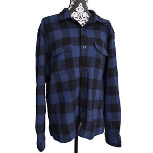 May include: A blue and black plaid flannel shirt with a button-down front, two chest pockets, and a collared neckline. The shirt is displayed on a mannequin. The fabric has a classic lumberjack style.