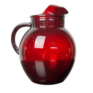 May include: A ruby red glass pitcher with a rounded body, a curved handle, and a spout with a lid. The pitcher is made of transparent glass, allowing light to pass through. The handle is a darker shade of red.