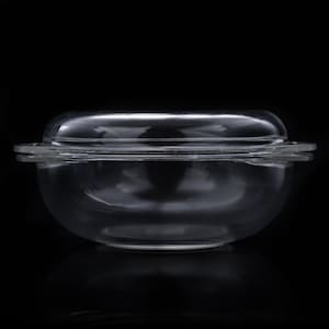 May include: A clear glass casserole dish with a matching lid. The round vessel features two integrated side handles and a smooth, transparent surface. The lid is slightly domed and fits securely over the top of the bowl, creating a cohesive unit.