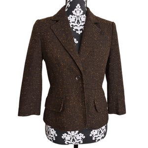 May include: A brown tweed blazer with a single-button closure and two front pockets. The blazer has three-quarter sleeves and is displayed on a mannequin. The fabric has flecks of gold and brown.