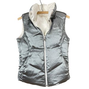 May include: A reversible silver puffer vest with a white interior, hanging on a wooden hanger. The vest has a full-length white zipper and a small Nike logo on the chest. The vest is designed with a high collar.