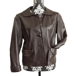 May include: A dark brown jacket featuring a subtle crocodile-embossed texture, a rounded collar, and three matching fabric-covered buttons down the front. The jacket includes two front patch pockets with curved flaps and long sleeves with cuffs.