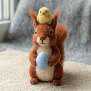 Könnte beinhalten: Eine handgefertigte Eichhörnchen-Figur aus Filz mit einem kleinen gelben Küken auf dem Kopf, das ein hellblaues Ei hält. Das Eichhörnchen hat braunes Fell, einen weißen Bauch und steht auf einer beigen Oberfläche.