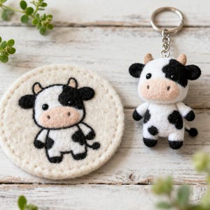 May include: A felt cow keychain and a matching coaster. The cow keychain is white with black spots, a black and white face, and a small tail. The coaster is round, cream-colored, and features a similar cow design.