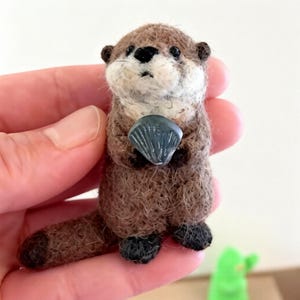 Op de afbeelding: Handgemaakte otterfiguur van bruine en crèmekleurige wol, met een kleine grijze schelp. De otter heeft zwarte ogen en een zwarte neus. De voeten zijn zwart. De achtergrond is licht. Een charmant decoratief item.