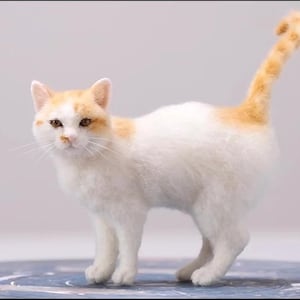 Op de afbeelding: Een naaldgevilte kattenfiguur met witte vacht en oranje vlekken op de kop en staart. De kat staat in een profielpositie met de staart omhoog, voorzien van gedetailleerde bruine ogen, een roze neus en een zacht, getextureerd uiterlijk.