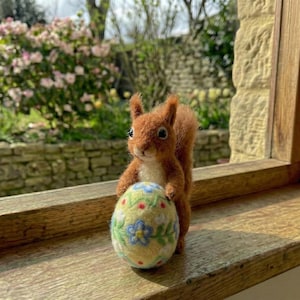 Könnte beinhalten: Eine handgefertigte Eichhörnchenfigur aus Filz, die ein dekoratives Osterei hält. Das Eichhörnchen ist braun mit weißem Bauch, und das Ei ist gelb mit blauen und roten Blumenmustern. Die Figur steht auf einer Holzfensterbank.
