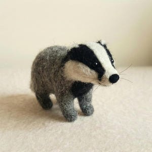 以下が含まれることがあります： 手作りのフェルト製のアナグマの置物。アナグマは灰色の体で、頭と顔に黒と白の模様があります。小さな黒い目と黒い鼻があります。置物は明るい色の表面に立っています。