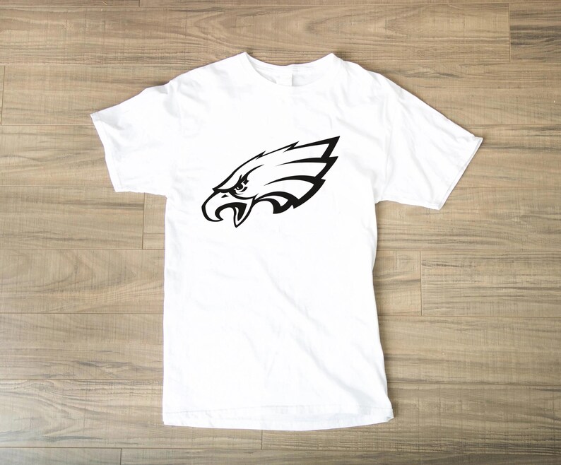 Pode incluir: Camiseta branca com um gr&aacute;fico preto da cabe&ccedil;a de uma &aacute;guia. O desenho da &aacute;guia &eacute; uma imagem estilizada com linhas n&iacute;tidas e um olho detalhado, contrastando com o tecido branco da camiseta.