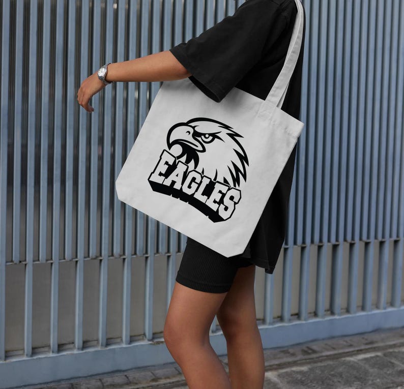 Pode incluir: Saco tote branco com um gr&aacute;fico preto da cabe&ccedil;a de uma &aacute;guia e a palavra "EAGLES" em letras grossas. O saco tem um design simples com al&ccedil;as longas e &eacute; usado contra um fundo azul e cinza.