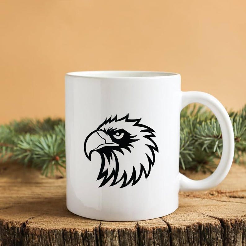 Pode incluir: Caneca de cer&acirc;mica branca com um desenho de cabe&ccedil;a de &aacute;guia preta. O gr&aacute;fico da &aacute;guia &eacute; uma ilustra&ccedil;&atilde;o detalhada com linhas n&iacute;tidas e um olhar focado. A caneca tem uma al&ccedil;a curva.
