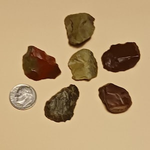 May include: An assortment of six polished stones in shades of red, brown, and green, arranged around a dime for size comparison. The stones have a smooth, natural texture and varying shapes, suggesting they are gemstones or decorative rocks.