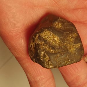 Puede incluir: Una piedra marrón oscura, de forma irregular, con inclusiones doradas, sostenida en una mano. La piedra tiene una textura rugosa y está parcialmente iluminada, revelando sus patrones naturales. Se ve un anillo plateado.