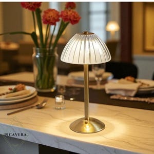 May include: A small, gold-coloured table lamp with a ribbed, white glass shade. The lamp is on a marble-effect surface, with a vase of red flowers in the background. The lamp is designed for ambient lighting.