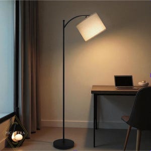 May include: A modern black arc floor lamp with a rectangular beige lampshade. The lamp is positioned near a window with a dark curtain. A desk with a laptop and chair are in the background.