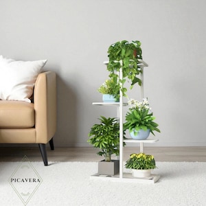 May include: A white, multi-tiered plant stand with several potted plants. The stand has a rectangular base and multiple shelves. The plants include green leafy plants and flowering plants in blue and white pots. The logo "Picavera" is at the bottom.
