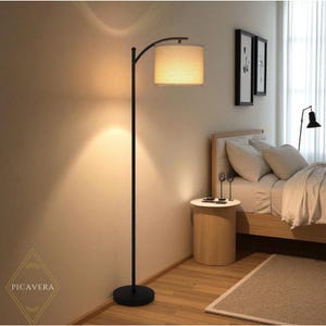 May include: A black arc floor lamp with a beige lampshade illuminates a bedroom. The lamp has a round base and a curved arm. The room features a bed, side table, and artwork. The overall aesthetic is modern and minimalist.