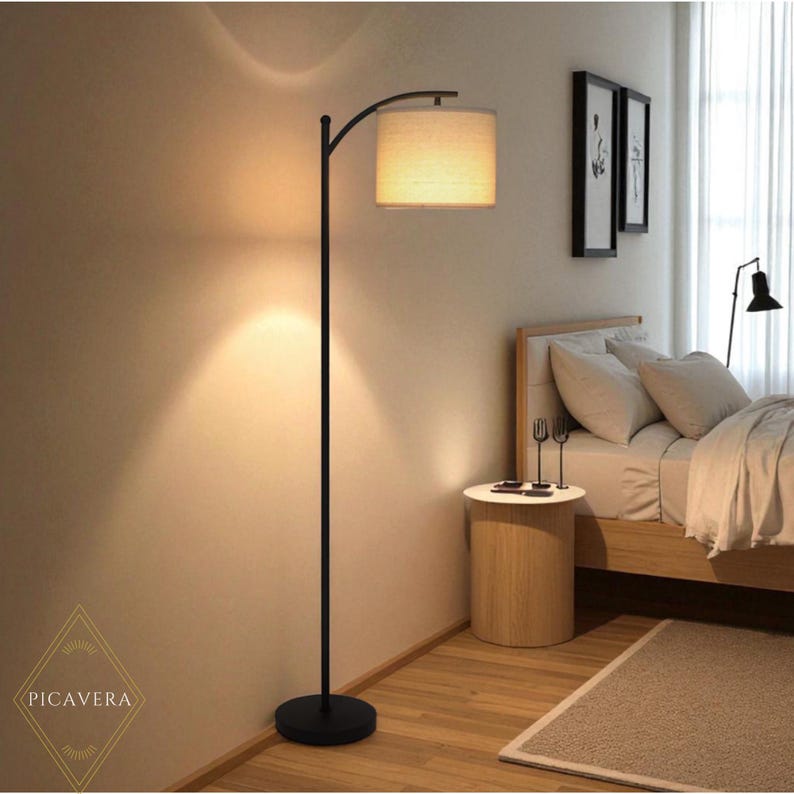 May include: A black arc floor lamp with a beige lampshade illuminates a bedroom. The lamp has a round base and a curved arm. The room features a bed, side table, and artwork. The overall aesthetic is modern and minimalist.
