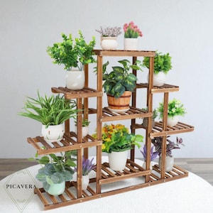 May include: A multi-level wooden plant stand displaying an assortment of potted plants and flowers. The stand is constructed from dark brown wood with a slatted design. The plants feature green foliage and vibrant flowers in white, brown, and light blue pots.