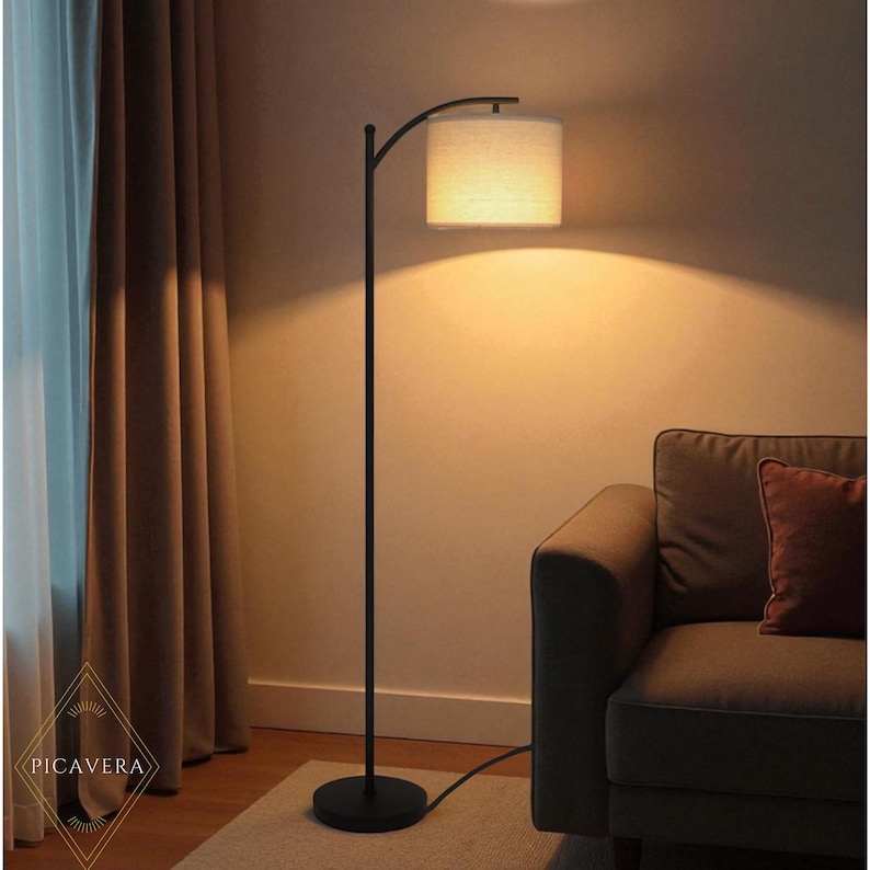 May include: A black, modern floor lamp with a curved arm and a cylindrical lampshade. The lampshade is a light beige color, and the lamp is positioned next to a sofa. The lamp's base is round.
