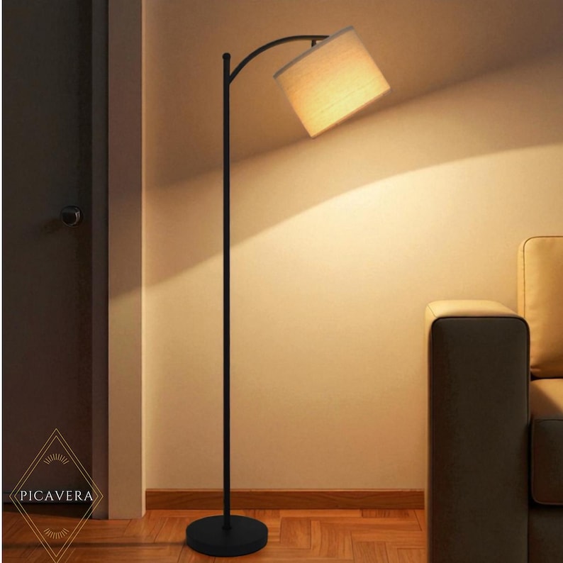 May include: A black floor lamp with a curved arm and a beige lampshade. The lamp has a round base and is positioned next to a wall and a sofa. The lampshade is angled downward, and the light illuminates the wall.