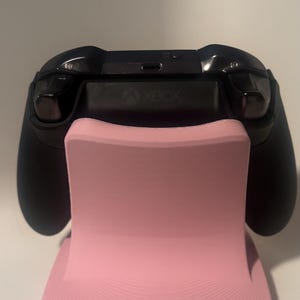 Puede incluir: Un mando Xbox negro descansa sobre un soporte rosa. El soporte tiene una forma curva y una base texturizada. El logotipo de Xbox es visible en el mando. El soporte est&aacute; dise&ntilde;ado para sostener el mando en posici&oacute;n vertical.