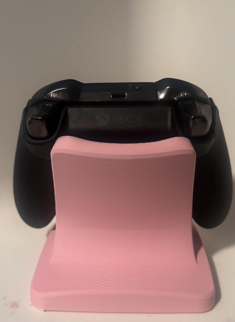 Puede incluir: Un mando Xbox negro descansa sobre un soporte rosa. El soporte tiene una forma curva y una base texturizada. El logotipo de Xbox es visible en el mando. El soporte est&aacute; dise&ntilde;ado para sostener el mando en posici&oacute;n vertical.