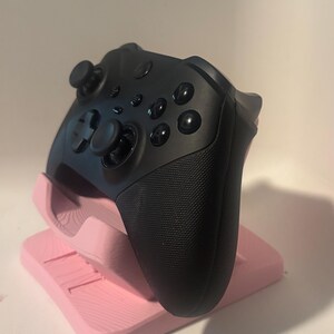 Puede incluir: Un mando de videojuegos negro descansa sobre un soporte rosa. El mando tiene varios botones, dos joysticks y agarres texturizados. El soporte es de color rosa claro y tiene un dise&ntilde;o en capas.