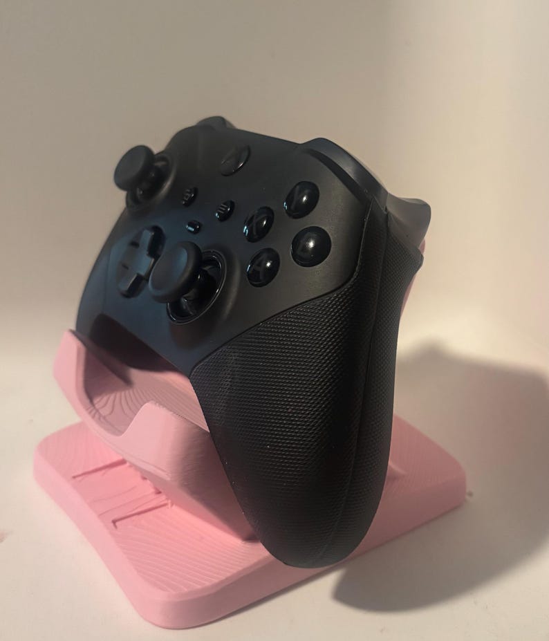 Puede incluir: Un mando de videojuegos negro descansa sobre un soporte rosa. El mando tiene varios botones, dos joysticks y agarres texturizados. El soporte es de color rosa claro y tiene un dise&ntilde;o en capas.