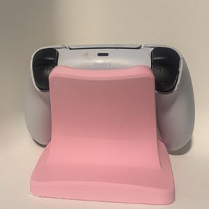 Puede incluir: Un soporte rosa para mando de videojuegos con un mando blanco. El soporte tiene una forma curva y una parte delantera texturizada. El mando tiene botones negros y se ve la palabra "SONY".