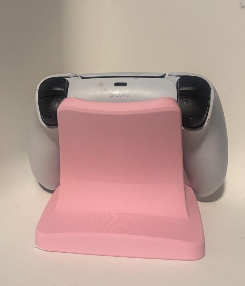 Puede incluir: Un soporte rosa para mando de videojuegos con un mando blanco. El soporte tiene una forma curva y una parte delantera texturizada. El mando tiene botones negros y se ve la palabra "SONY".