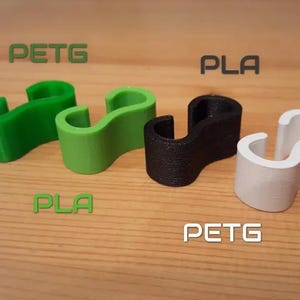 Peut inclure: Quatre crochets en forme de S imprimés en 3D, de couleurs verte, noire et blanche, disposés sur une surface en bois. Les crochets portent les inscriptions "PETG" et "PLA".