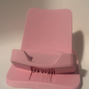 Puede incluir: Soporte para tel&eacute;fono rosa liso con base texturizada y respaldo. El soporte tiene un borde curvo para sujetar el tel&eacute;fono y peque&ntilde;os soportes en la parte inferior. El soporte est&aacute; hecho de un material s&oacute;lido.