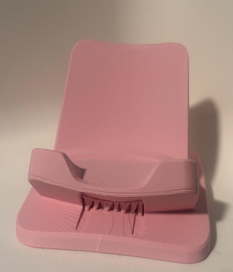 Puede incluir: Soporte para tel&eacute;fono rosa liso con base texturizada y respaldo. El soporte tiene un borde curvo para sujetar el tel&eacute;fono y peque&ntilde;os soportes en la parte inferior. El soporte est&aacute; hecho de un material s&oacute;lido.
